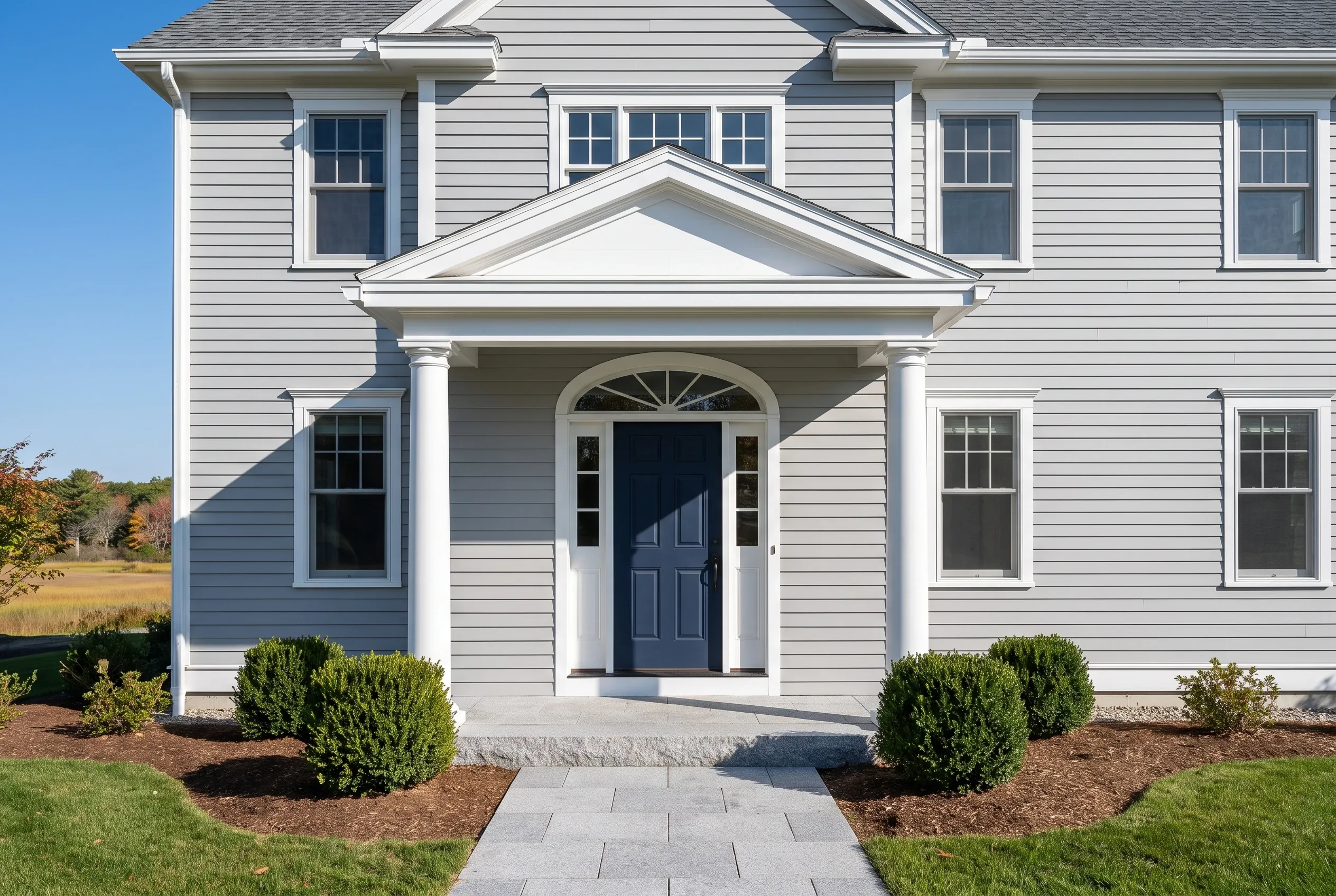 White Colonial Home in Scarborough, ME: Two-story white vinyl-sided colonial with gray roof, white trim, dark blue front door, columned portico, and brick paver walkway flanked by landscaping.